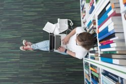 Studentin sitzt in einer Bibliothek an einem Bücherregal und arbeitet mit einem Laptop